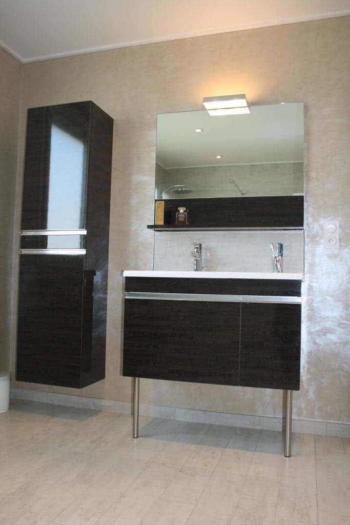 Meuble-lavabo moderne avec miroir, placards foncés, lavabo et armoire de rangement haute, le tout adossé à un mur carrelé beige.