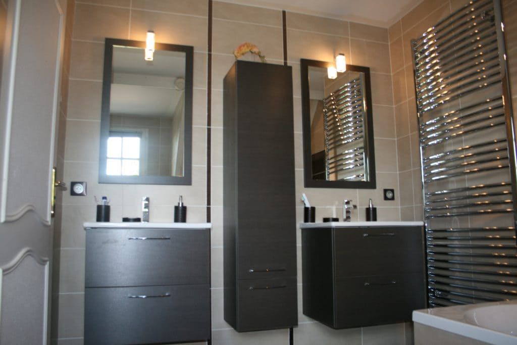 Salle de bains moderne avec deux lavabos, de grands miroirs, des meubles noirs, des appliques murales et un store à la fenêtre de droite.
