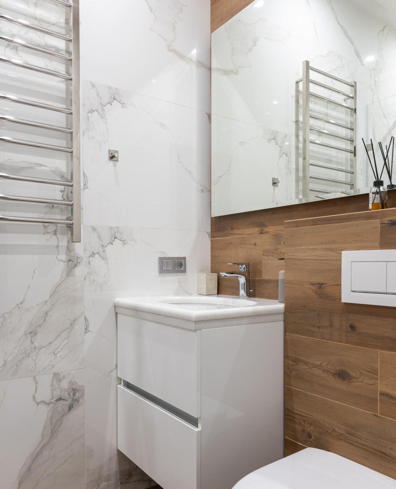 Meuble-lavabo moderne avec miroir, murs aspect marbre blanc, panneau décoratif en bois et sèche-serviettes chromés.
