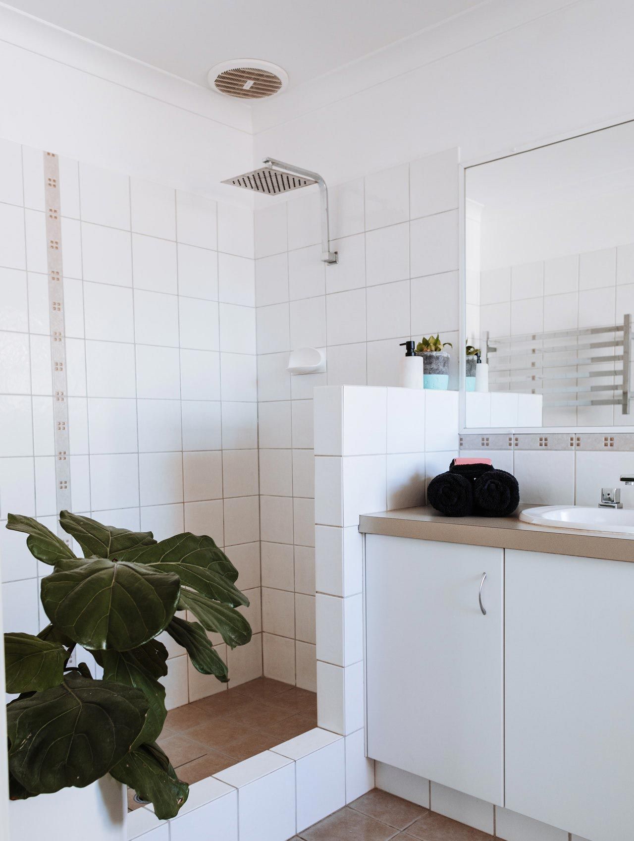 Salle de bains lumineuse et blanche avec douche carrelée, meuble-lavabo, miroir et grande plante verte.