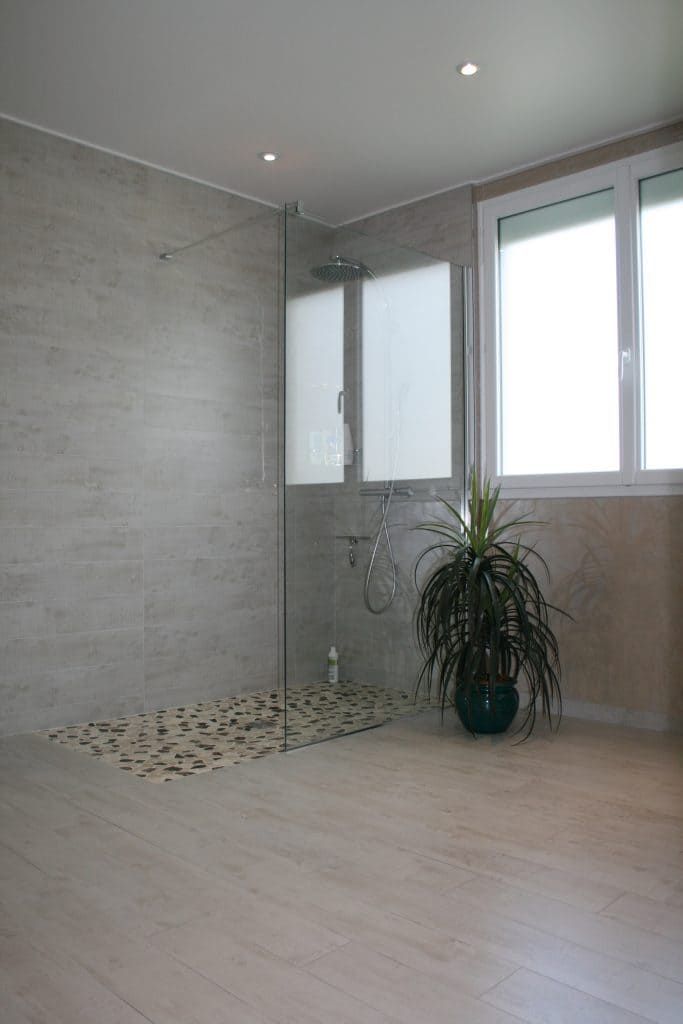 Salle de bains moderne et minimaliste avec murs carrelés gris, grandes fenêtres, sol de douche en galets et plante en pot.
