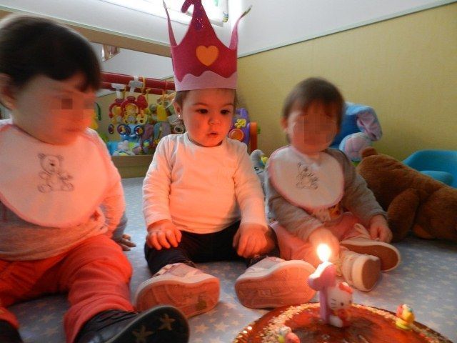 Tres bebés con baberos y coronas están sentados frente a un pastel de cumpleaños.