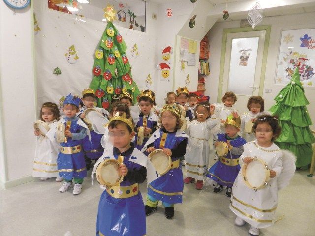 Un grupo de niños vestidos de ángeles sostienen una pandereta frente a un árbol de Navidad.