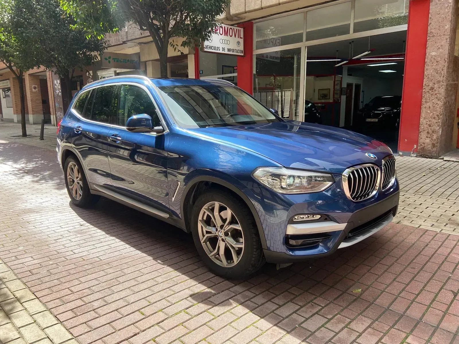 Un BMW X3 azul está estacionado en una acera de ladrillos frente a un concesionario de automóviles.