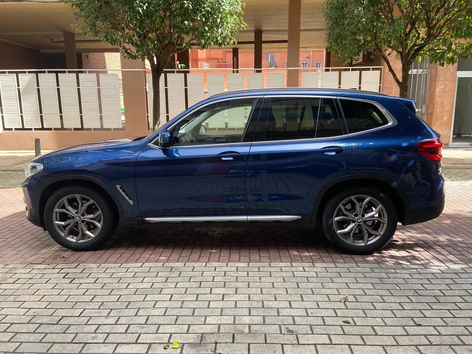 Un BMW X3 azul está estacionado en una acera de ladrillos frente a un edificio.