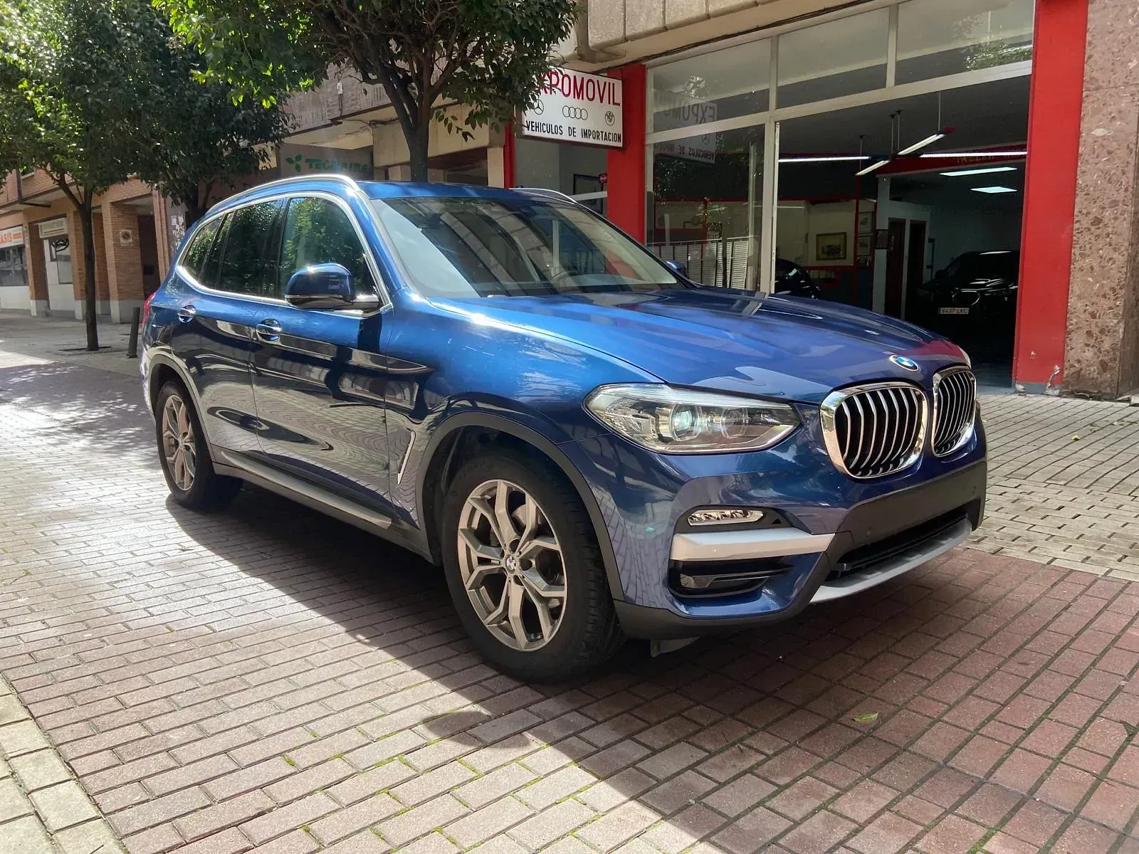 Un BMW X3 azul está estacionado en una acera de ladrillos frente a un concesionario de automóviles.