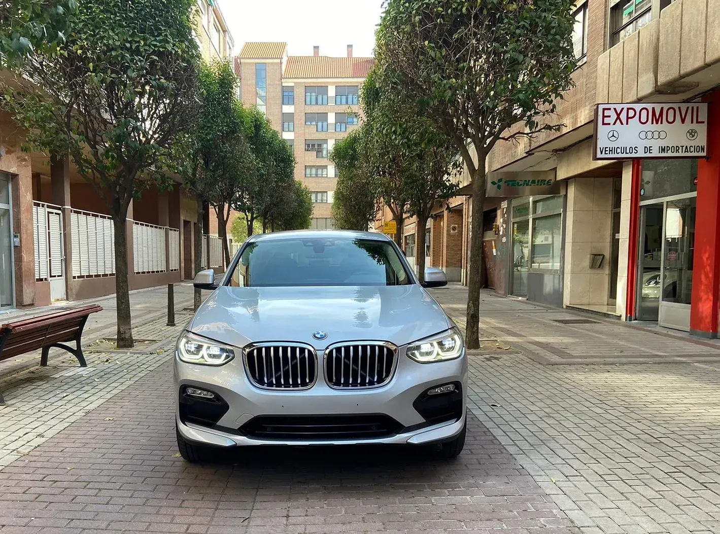 Un BMW plateado está estacionado en una acera de ladrillos frente a un edificio.