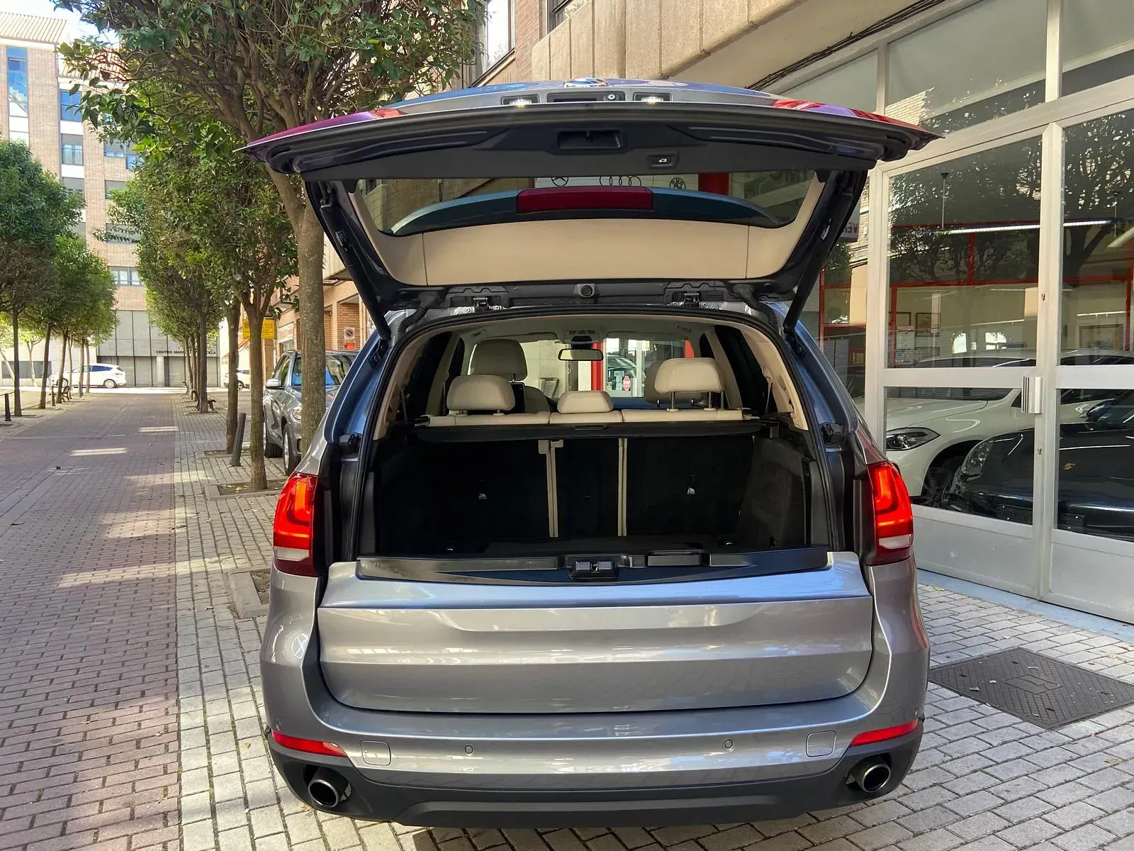 La parte trasera de un BMW X5 con el maletero abierto está estacionada sobre una acera de ladrillos.
