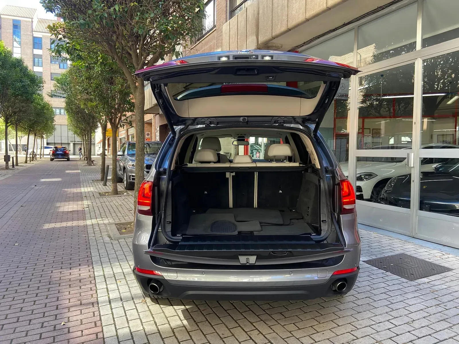 La parte trasera de un BMW X5 con el maletero abierto está estacionada sobre una acera de ladrillos.