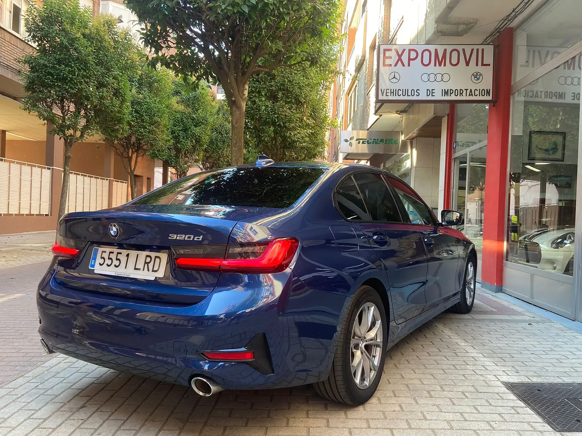 Un BMW Serie 3 azul está estacionado frente a un concesionario de automóviles.