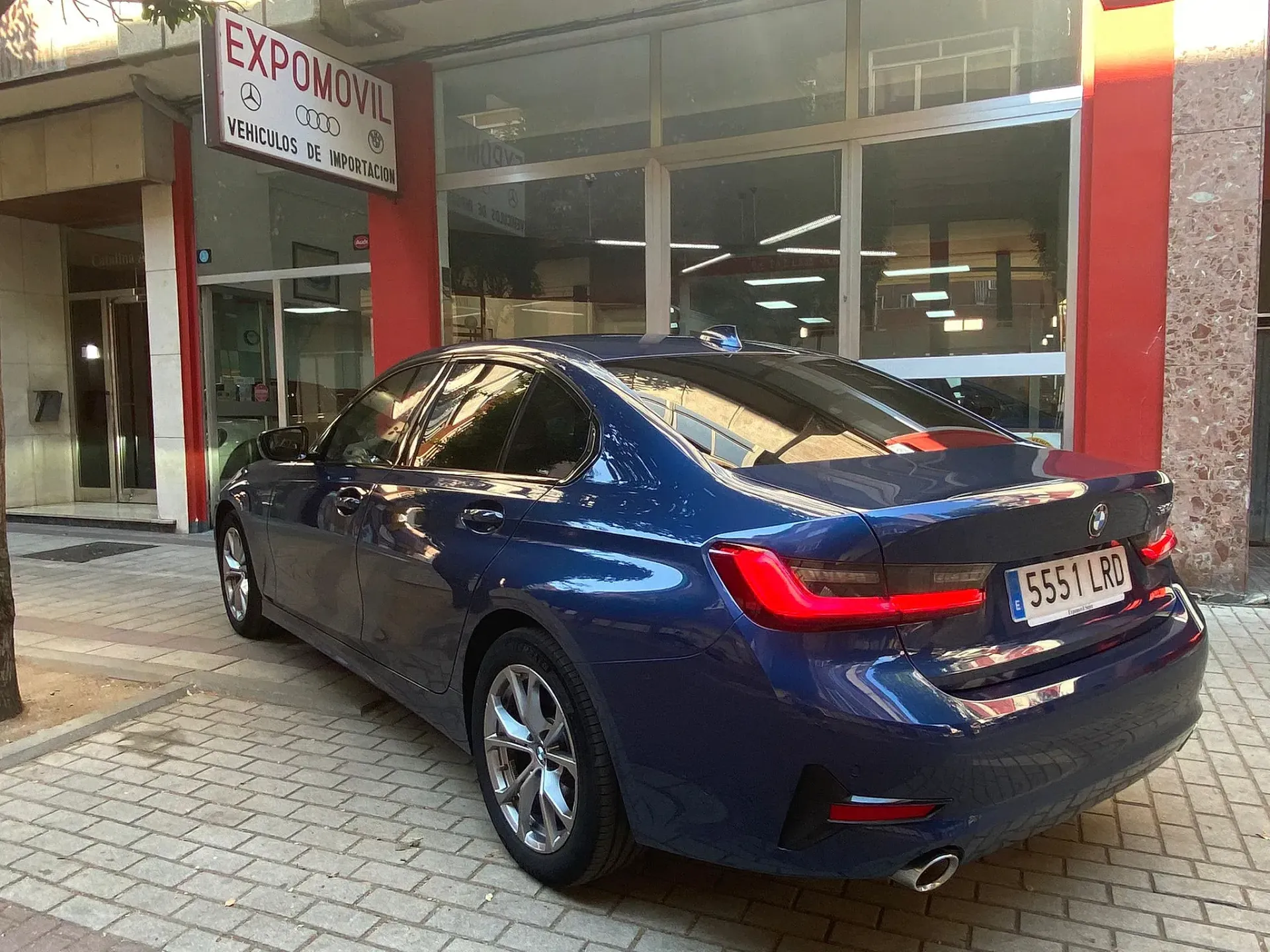 Un BMW Serie 3 azul está estacionado frente a un concesionario de automóviles.