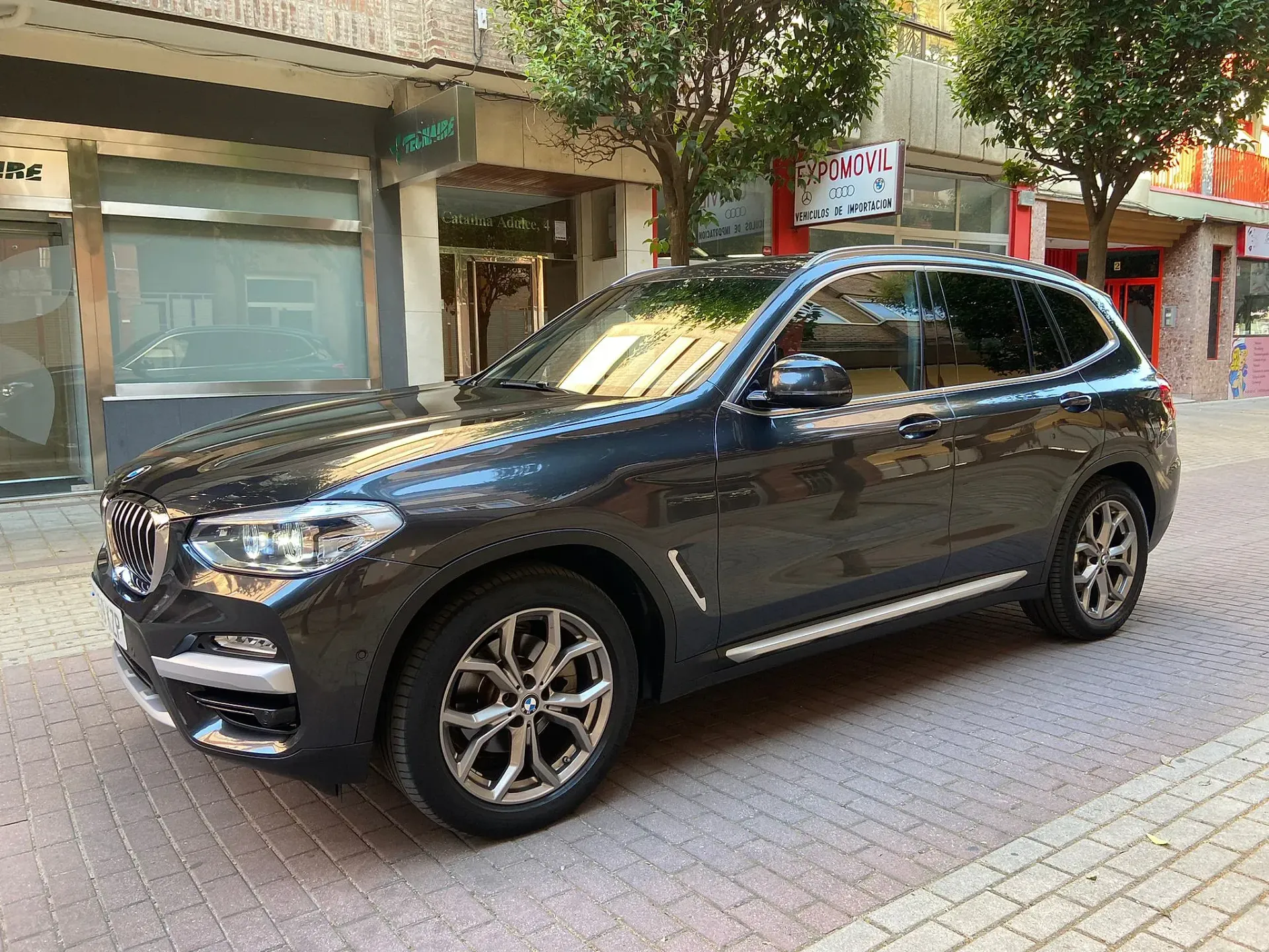 Un BMW X3 negro está estacionado en una acera de ladrillos frente a un edificio.