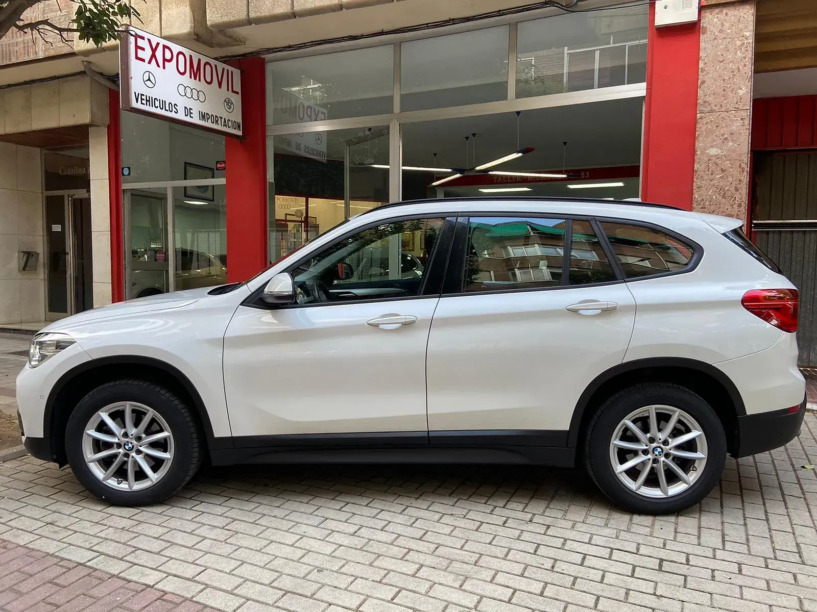 Un BMW X1 blanco está estacionado frente a un concesionario de automóviles.
