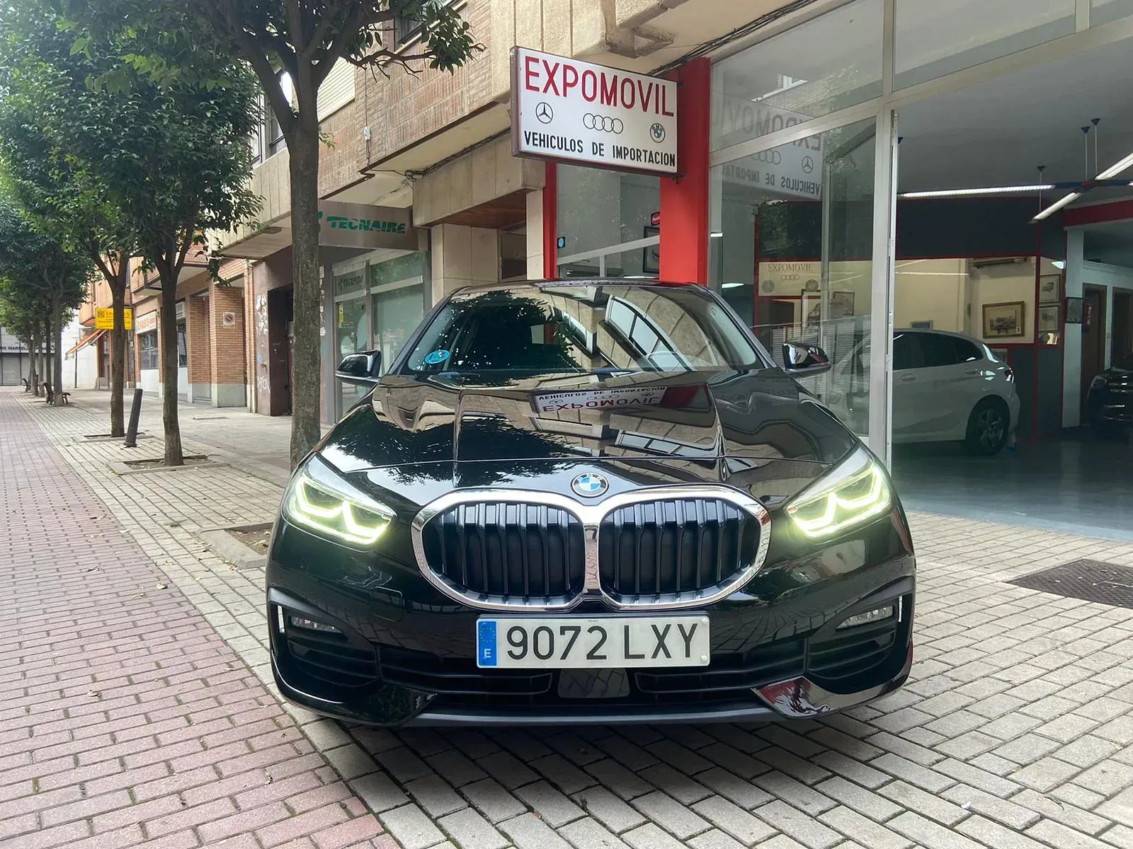 Un BMW negro está estacionado en una acera de ladrillos frente a un concesionario de automóviles.