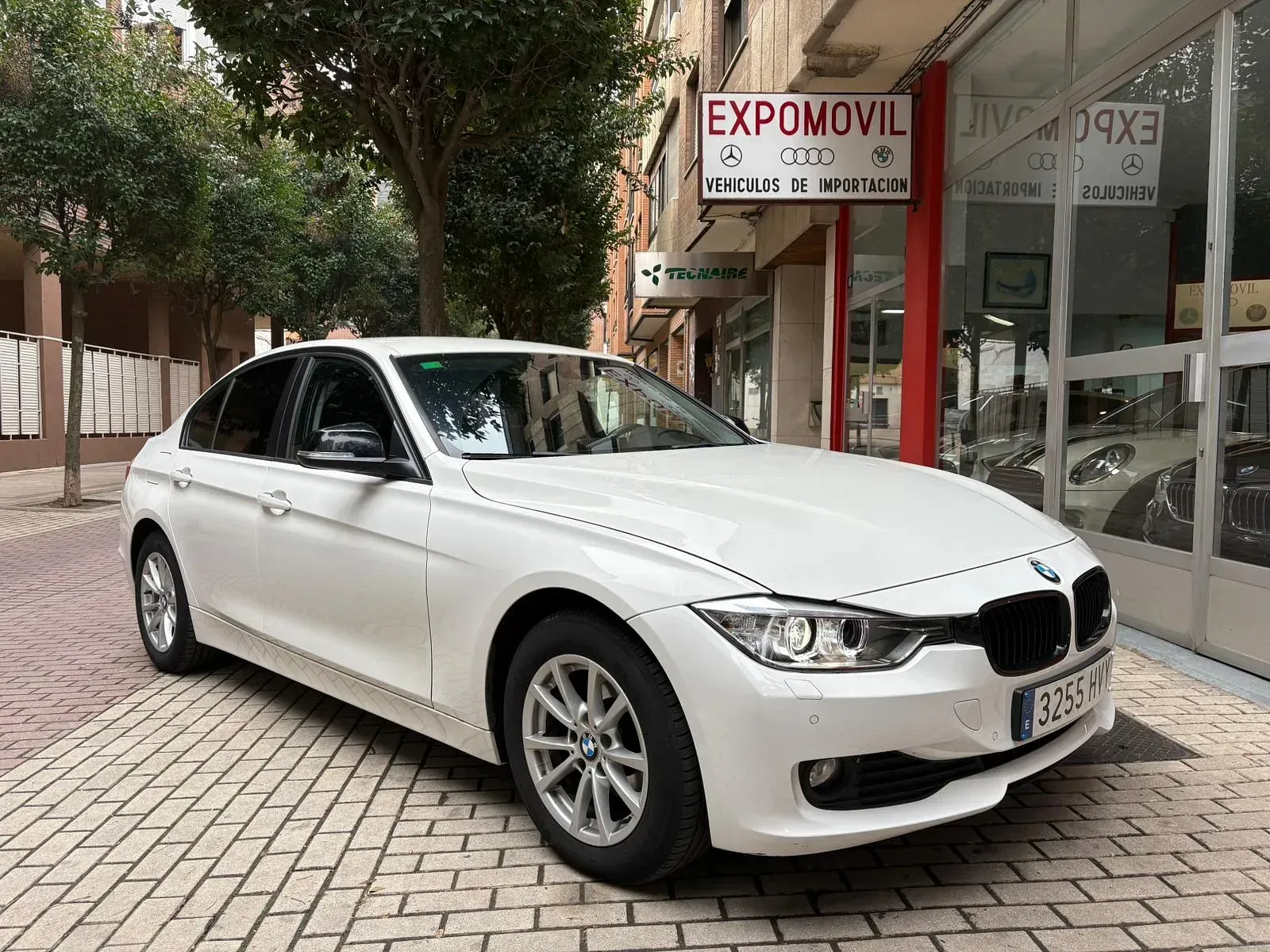 Un BMW Serie 3 blanco está estacionado frente a un concesionario de automóviles.