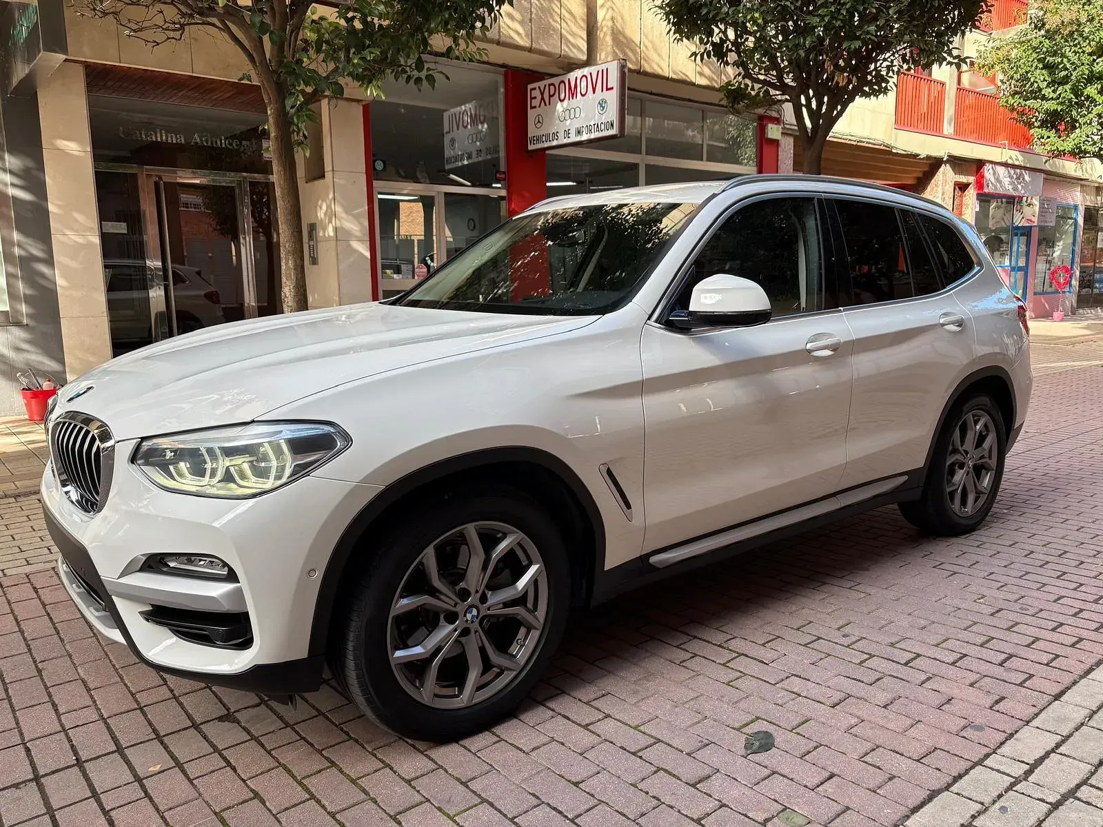Un BMW X3 blanco está estacionado en una acera de ladrillo frente a un edificio.