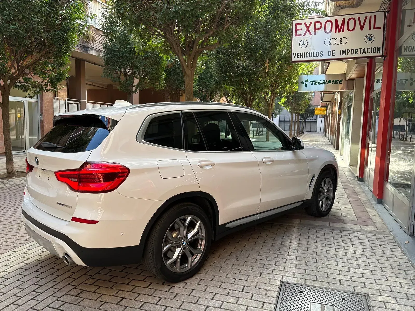 Una camioneta blanca está estacionada en una acera de ladrillos frente a un edificio.