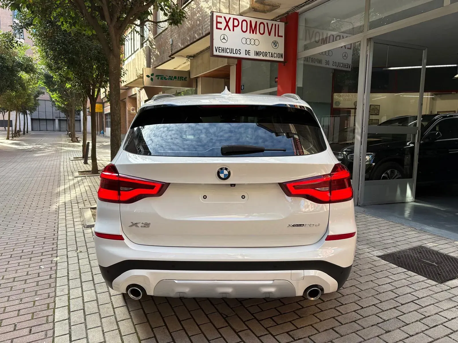 Un BMW X3 blanco está estacionado en una acera de ladrillos frente a un concesionario de automóviles.