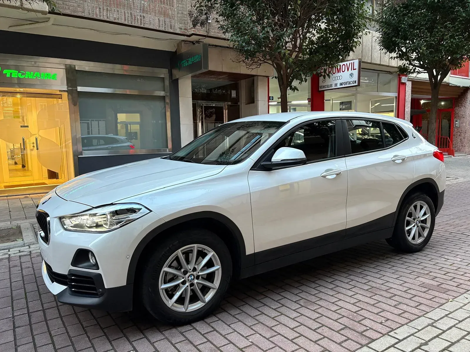 Un BMW X2 blanco está estacionado en una acera de ladrillos frente a un edificio.