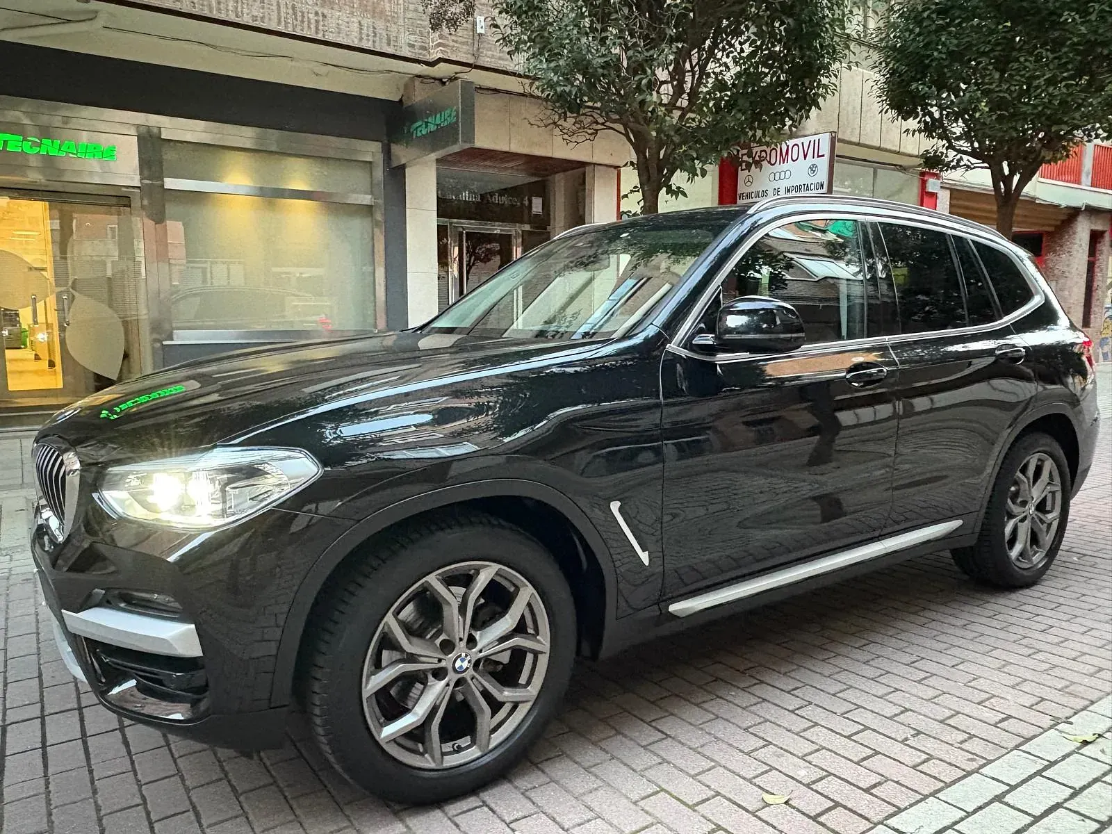Un BMW X3 negro está estacionado en una acera de ladrillos frente a un edificio.