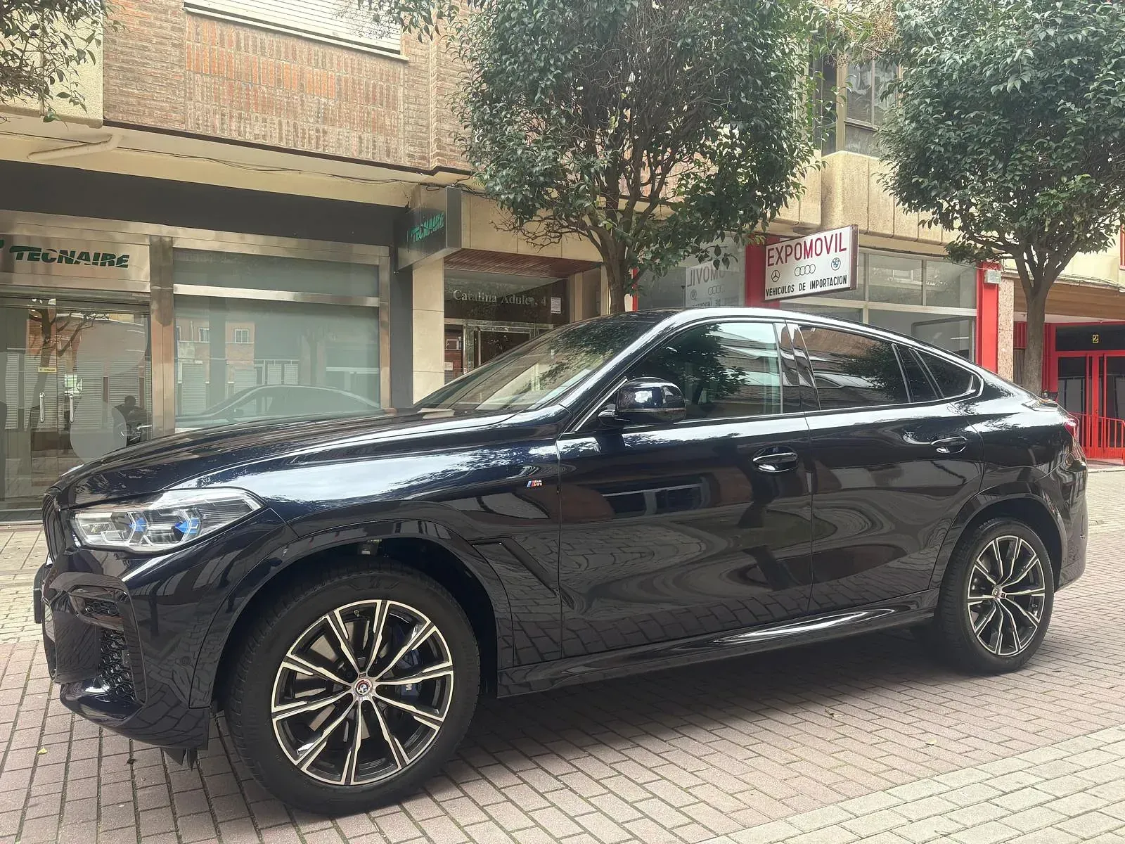 Un BMW X6 negro está estacionado en una acera de ladrillos frente a un edificio.