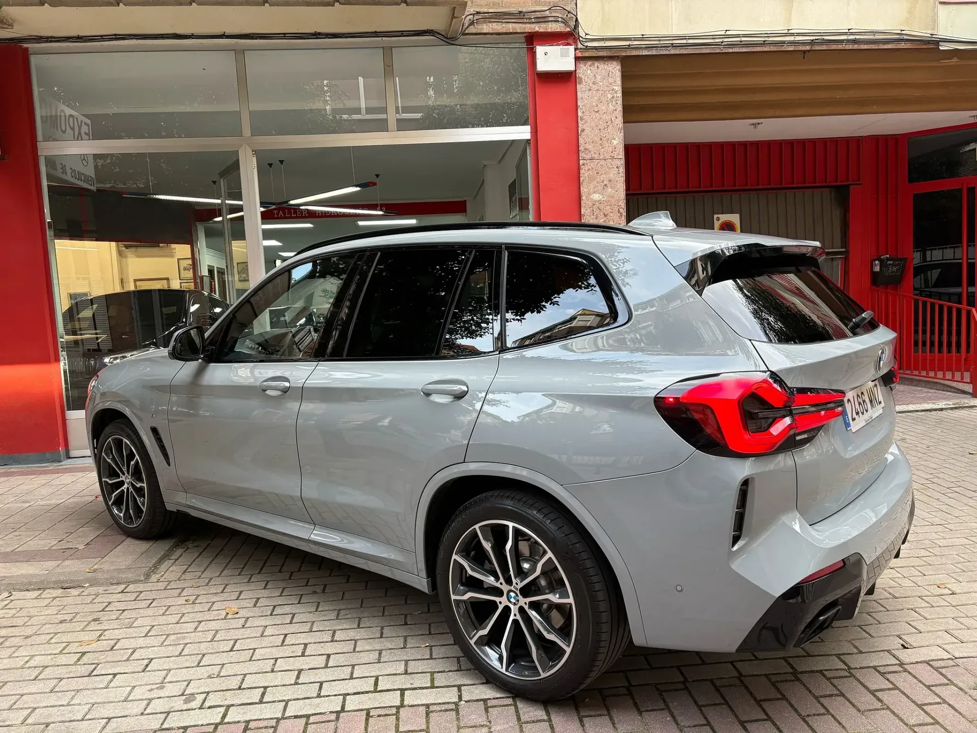 Un BMW X3 gris está estacionado frente a un edificio rojo.