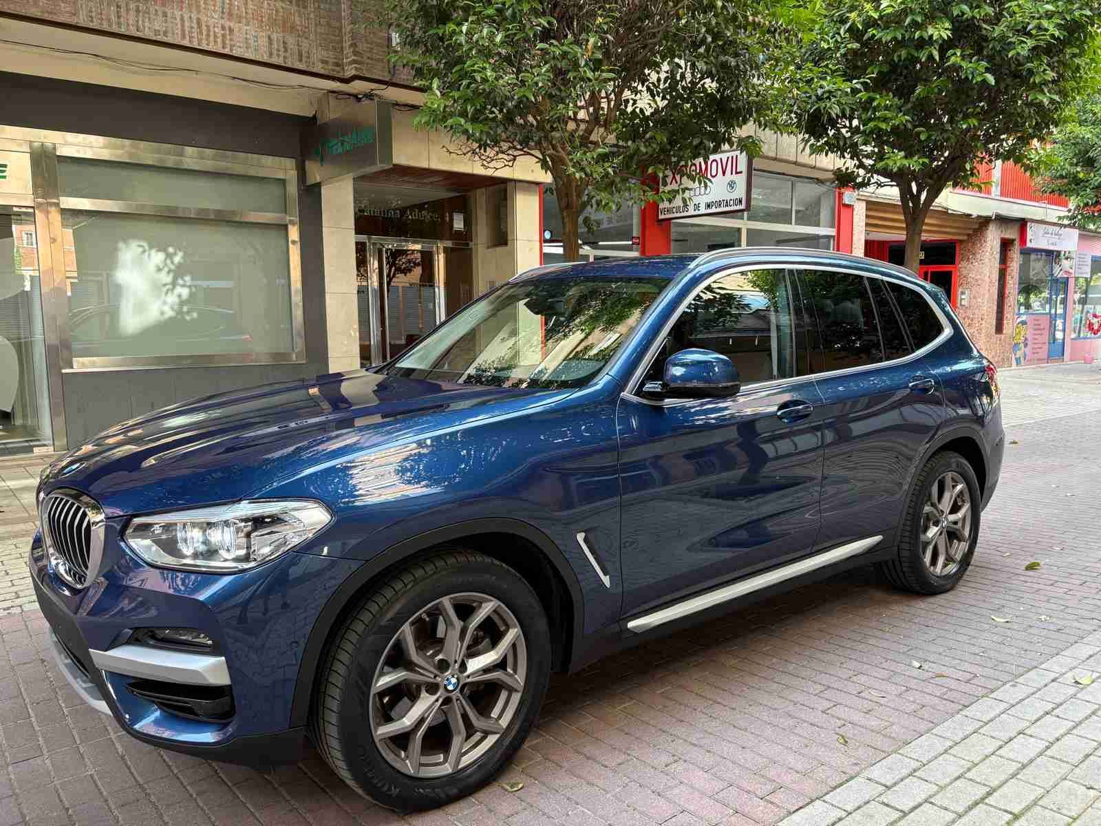 Un BMW X3 azul está estacionado en una acera de ladrillos frente a un edificio.