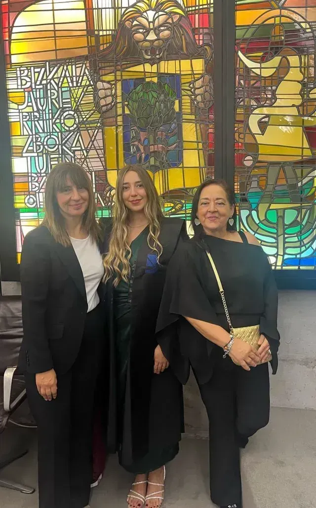 Tres personas están de pie una al lado de la otra, sonriendo frente a una colorida vidriera decorada con dibujos.