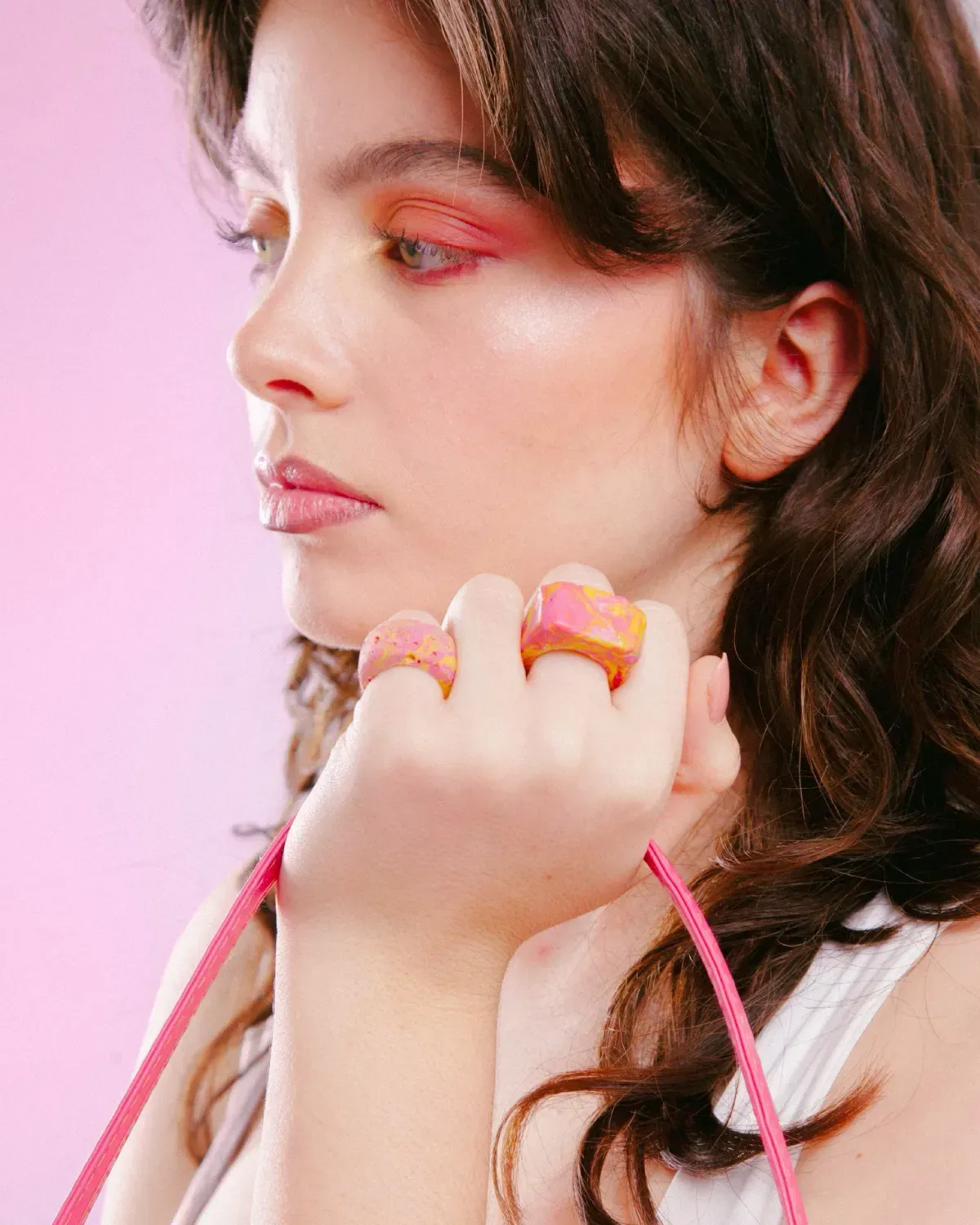 Retrato de perfil de una persona con maquillaje de ojos rosa, sosteniendo un anillo colorido sobre un fondo rosa pastel.