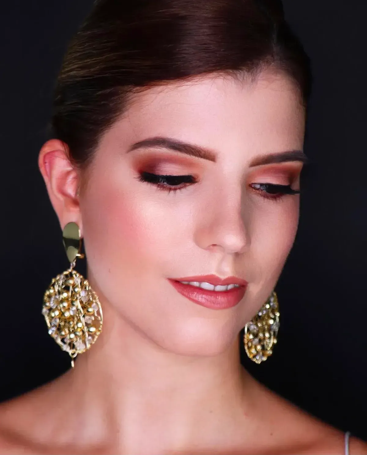 Retrato de una mujer con los ojos cerrados, luciendo pendientes de oro y maquillaje suave sobre un fondo oscuro.