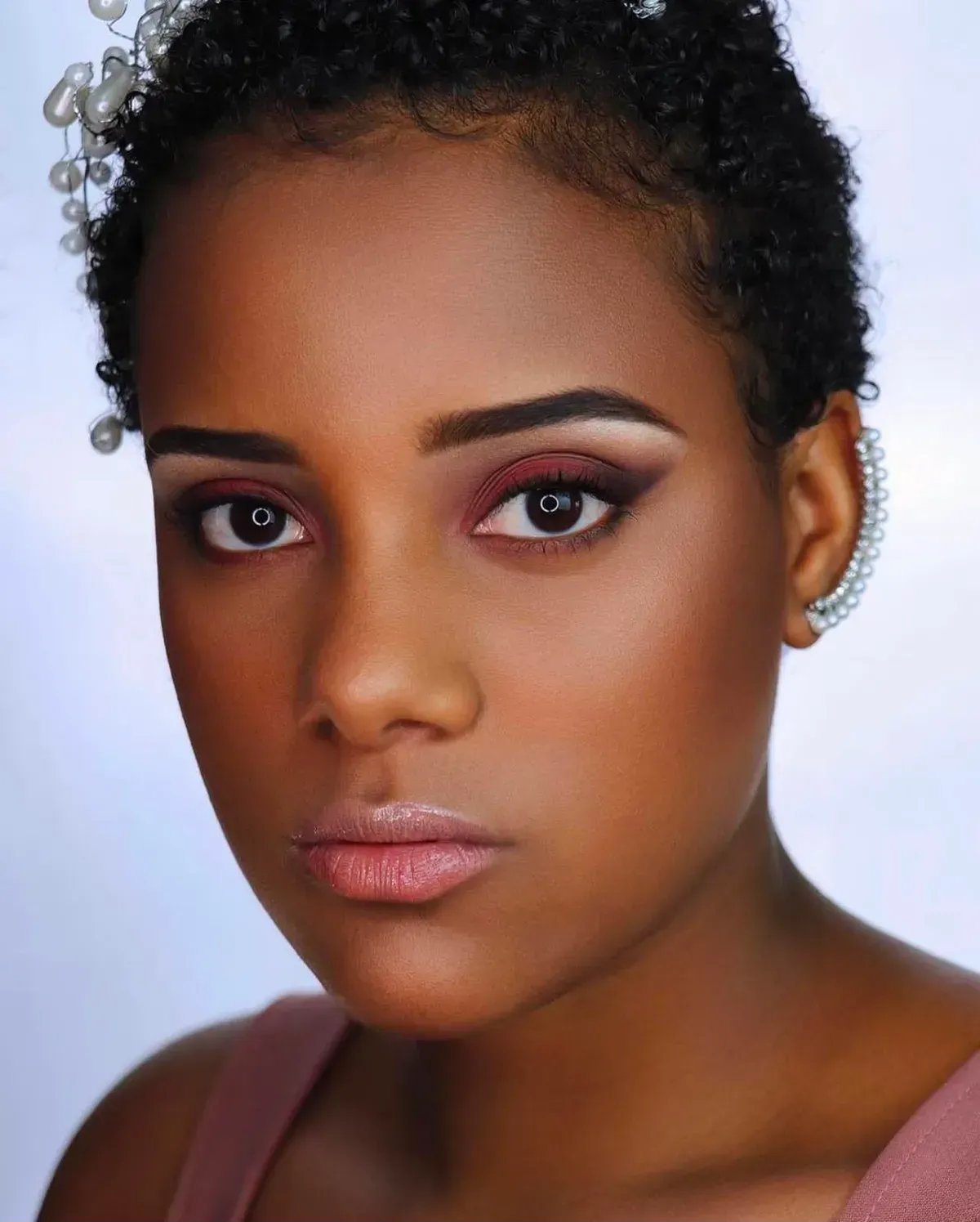 Retrato en primer plano de una persona con cabello corto y rizado, expresión neutra y maquillaje suave sobre un fondo claro.