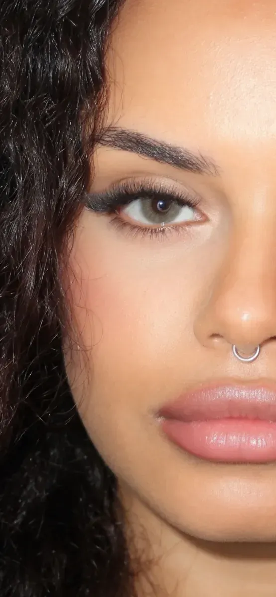 Primer plano de un rostro con cabello oscuro rizado, delineador de ojos alado, labios rosados ​​y un piercing