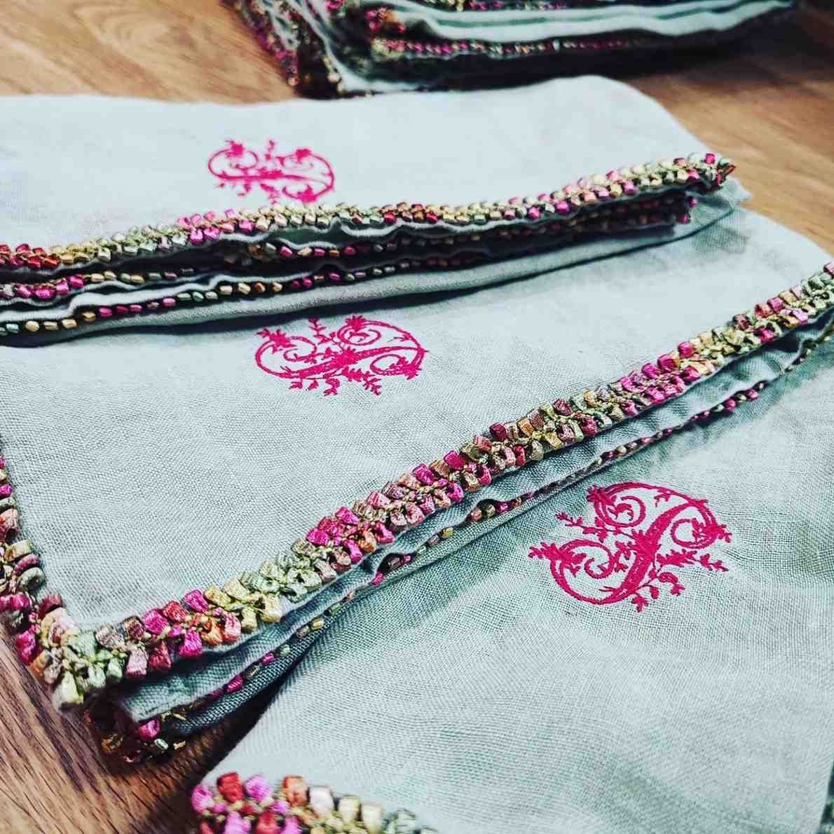 Servilletas apiladas de tela azul pálido con logotipos bordados en rosa fucsia y un borde decorativo.