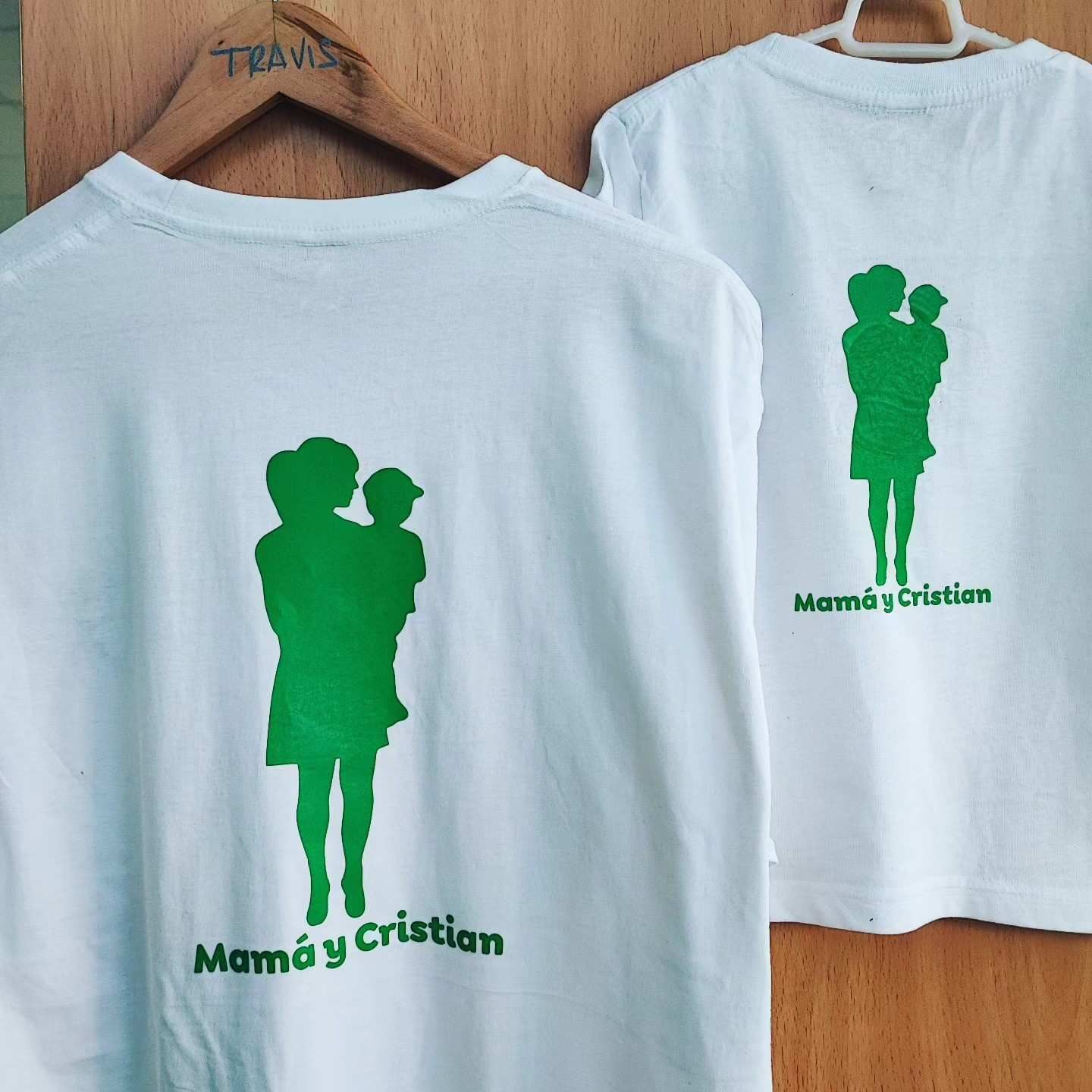 Dos camisetas blancas colgadas sobre un fondo de madera, cada una con la silueta verde de una persona que sostiene a un niño.