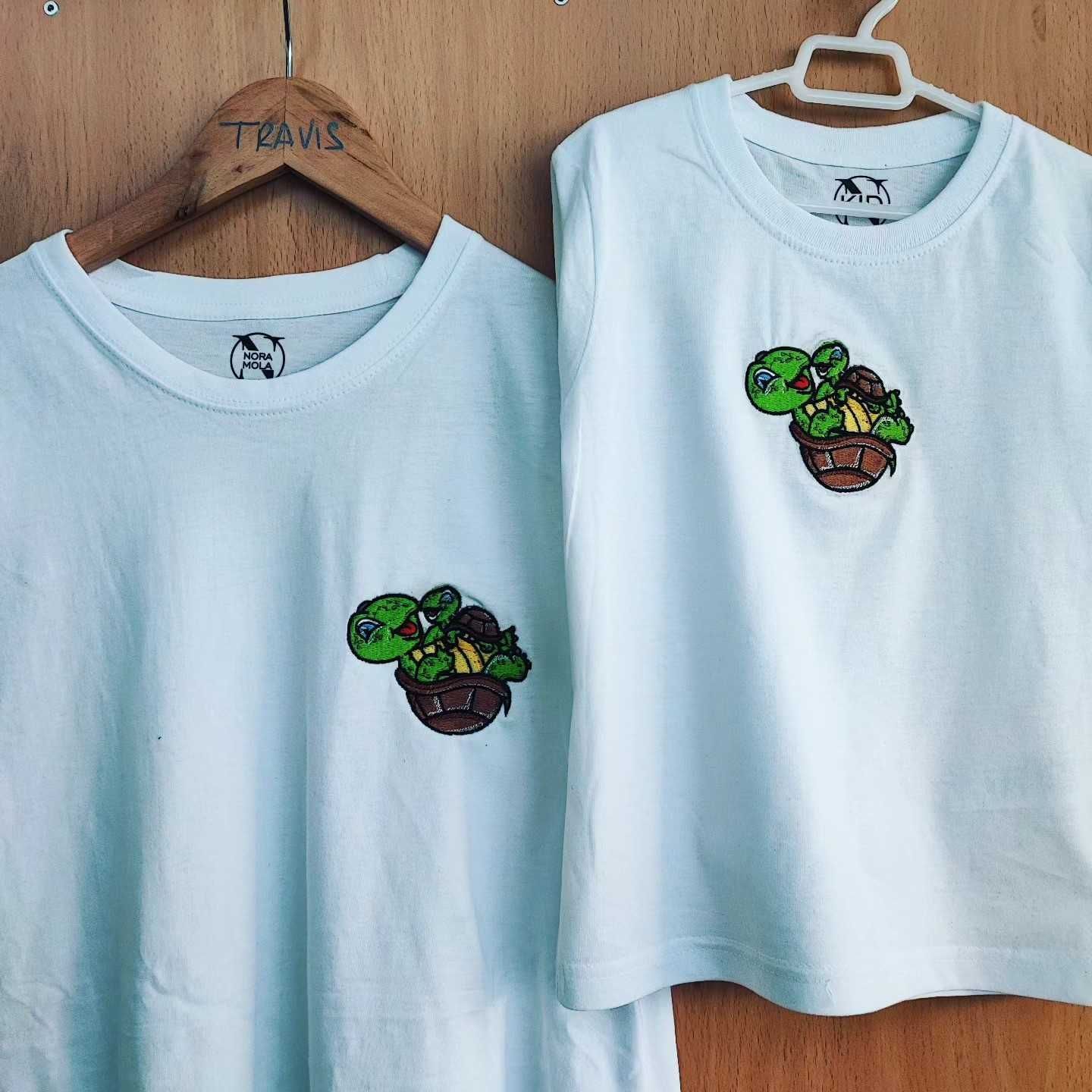 Dos camisetas blancas colgadas en perchas de madera, cada una con un diseño bordado de una tortuga de dibujos animados.