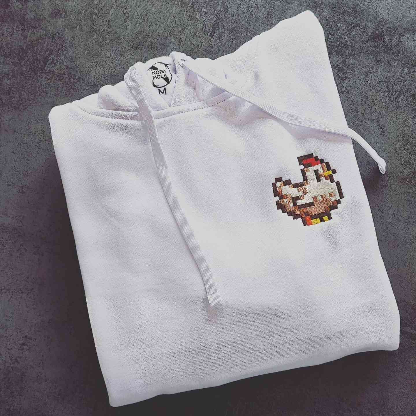 Una sudadera blanca doblada con un pequeño pollo estilo pixel art bordado en el pecho.