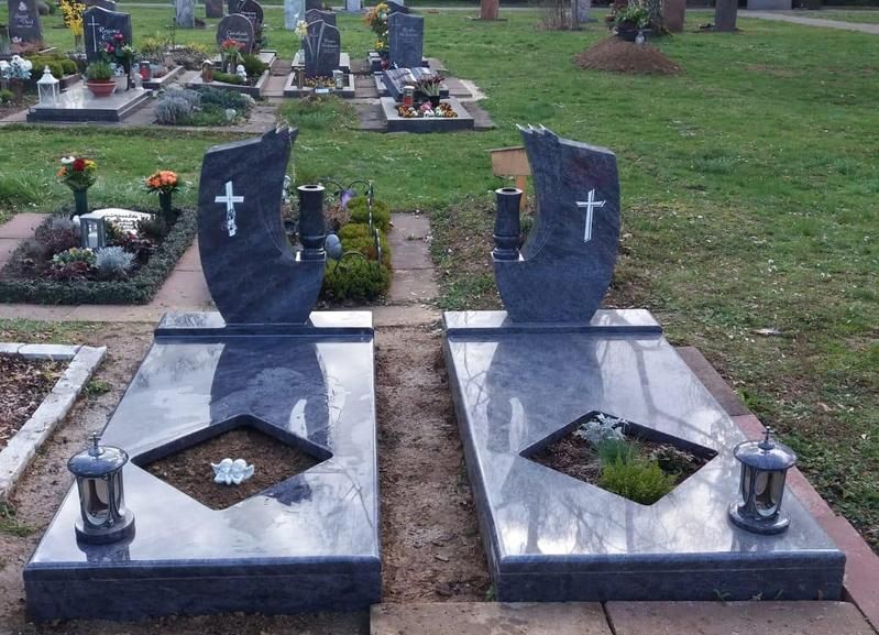 Zwei schwarze Granitgrabsteine ​​mit weißen Kreuzen auf einem Friedhof, jeder mit einem kleinen Blumenbeet und einer Laterne.