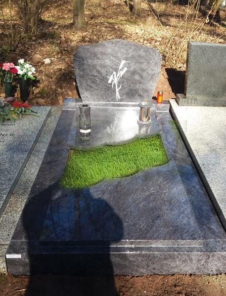Ein Granitgrabstein mit einem grünen Moosfleck und zwei Kerzen auf einem Friedhof, in der Nähe stehen Blumen.
