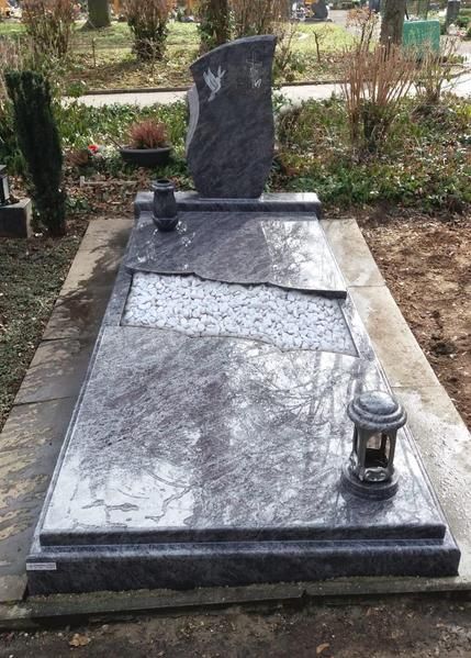 Grabmal aus schwarzem Granit mit Laterne in einem Friedhofsgarten mit Sträuchern.