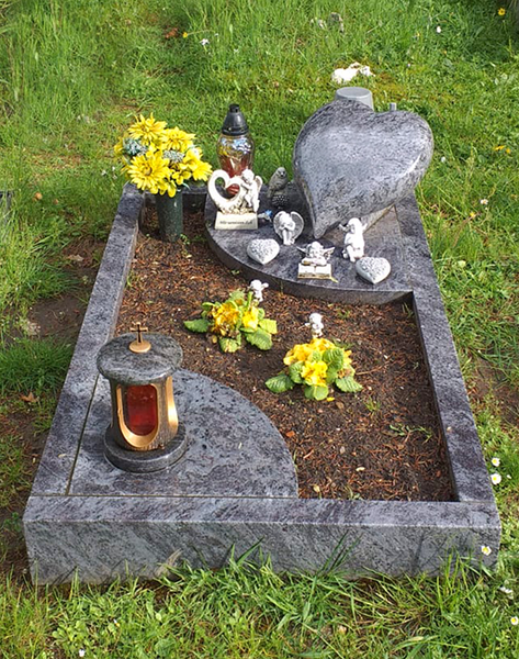 Grabstein-Gedenkstätte mit grauem, herzförmigem Stein, gelben Blumen, Kerzen und einer Laterne auf Rindenmulch im Gras