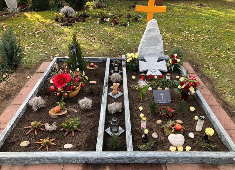 Ein mit Blumen, Kerzen und einem weißen, kreuzförmigen Grabstein geschmücktes Grab in einem Gartenbereich