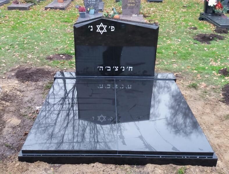 Schwarzer, polierter Grabstein mit hebräischer Inschrift auf einem Friedhof, der kahle Bäume spiegelt.