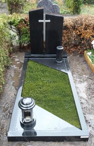 Schwarzer Grabstein mit weißem Kreuz und poliertem Grab, umgeben von Gras und Laternen auf einem Friedhof