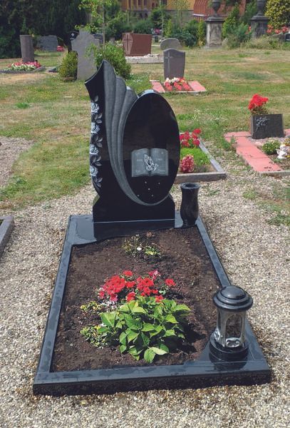 Schwarzer Gedenkgrabstein mit ovalem Porträt, roten Blumen und Laterne auf einem Friedhofsplatz