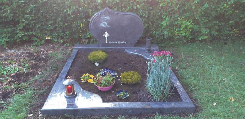 Grabstein mit Blumen und Kerzen auf einem kleinen, ordentlich bepflanzten Gedenkplatz im Freien
