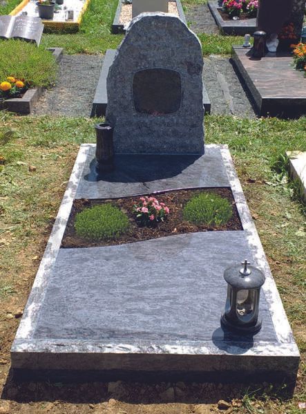 Grabstein mit dunklem Steinmarkierungsstein und Laterne auf einem von Gras und Blumen umgebenen Friedhofsplatz