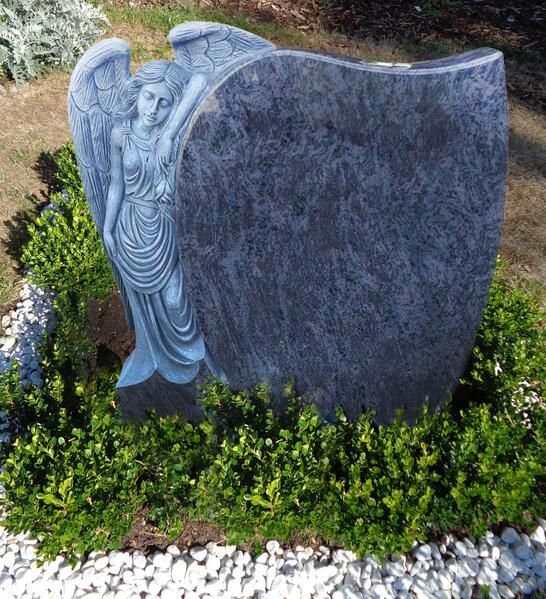 Steindenkmal mit einem geschnitzten Engel und einer leeren, dunklen Gedenktafel in einem Gartenbeet