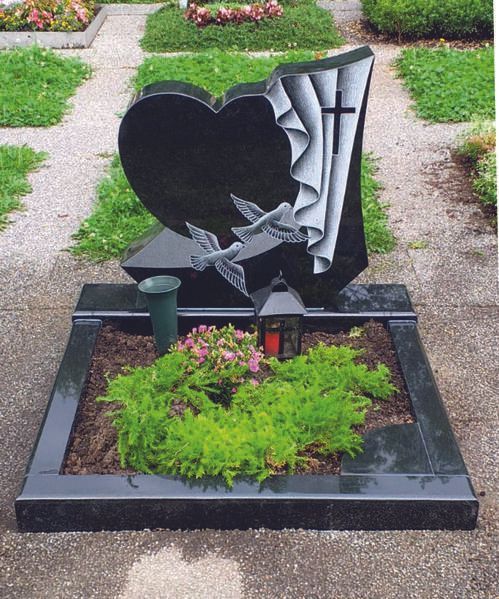 Schwarzer herzförmiger Grabstein mit silberner Kreuzskulptur in einem Blumen- und Grünbeet auf dem Friedhof