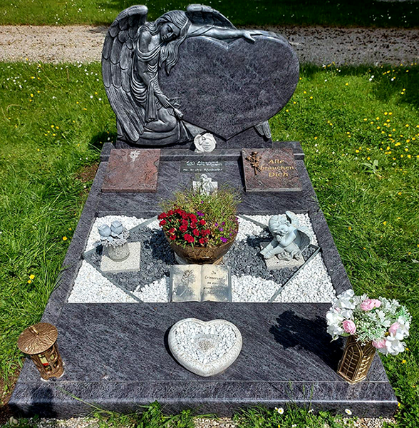 Ein mit Blumen geschmückter Grabstein mit einer herzförmigen Skulptur und gravierten Gedenktafeln auf einem Friedhof