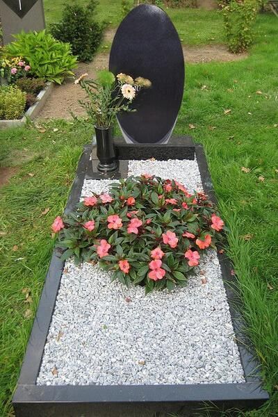 Grabdenkmal mit ovalem schwarzem Grabstein, Blumen und rosa Blüten auf weißem Kies